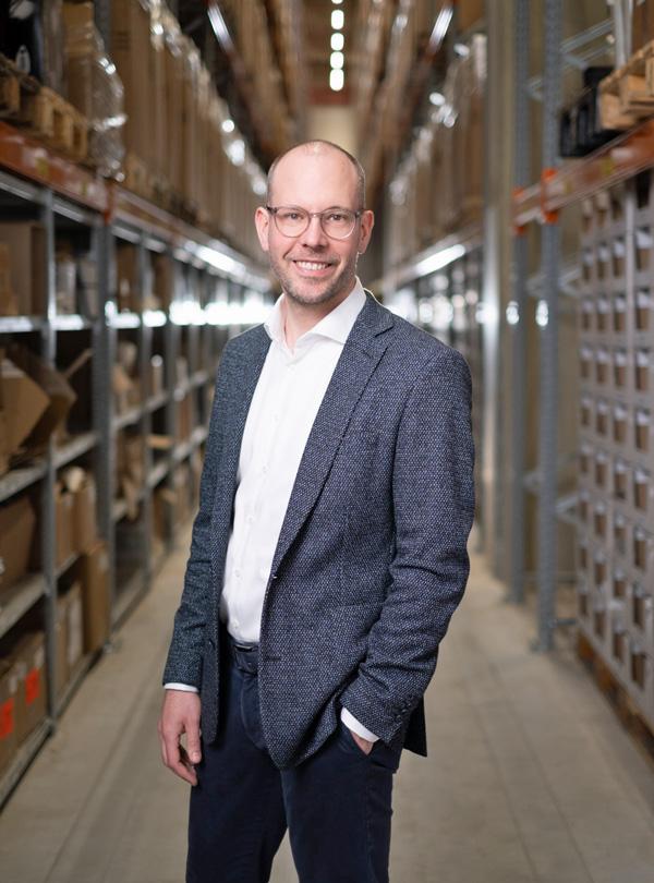 Niclas Meyer-Schuchardt, CFO atrikom, steht in einem Lagerhaus zwischen Regalen mit Kartons und Paketen, gekleidet in einem Blazer und Hemd.