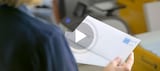 Person hält einen weißen Umschlag neben einem Bürodrucker; darüber liegt ein Wiedergabesymbol.