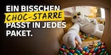 Ein cooler Osterhase mit Sonnenbrille liegt auf einem Sofa und bedient sich aus einem Paket mit Schoko-Eiern. Die andere Hand liegt auf seinem rundlichen Bauch. Schriftzug: Ein bisschen Schockstarre passt in jedes Paket.