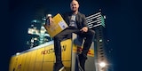Jürgen Vogel sitzt auf Packstation mit Paket im Arm.