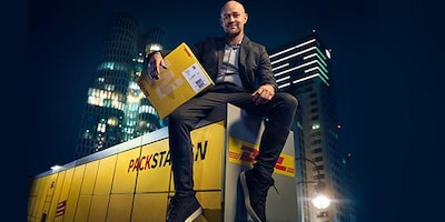 Jürgen Vogel sitzt auf Packstation mit Paket im Arm.